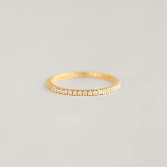 A delicate Gold band ring with small diamonds encircling it is placed on a soft, neutral background, exuding elegance and simplicity.