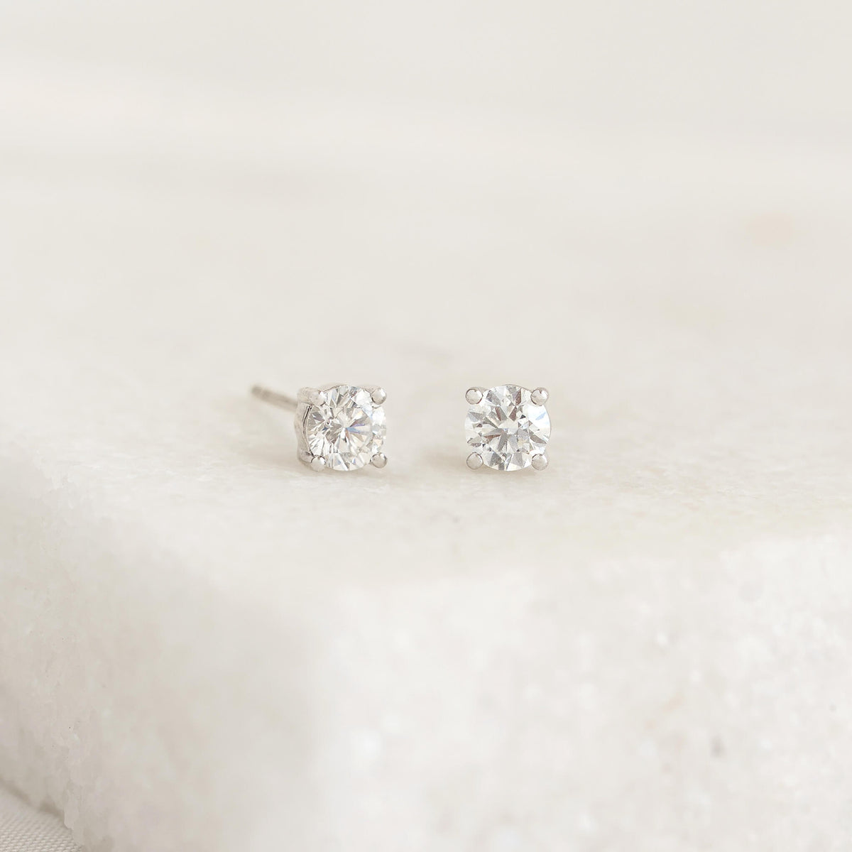 Two diamond stud earrings with White Gold settings are placed on a white textured surface. The image conveys elegance and simplicity.