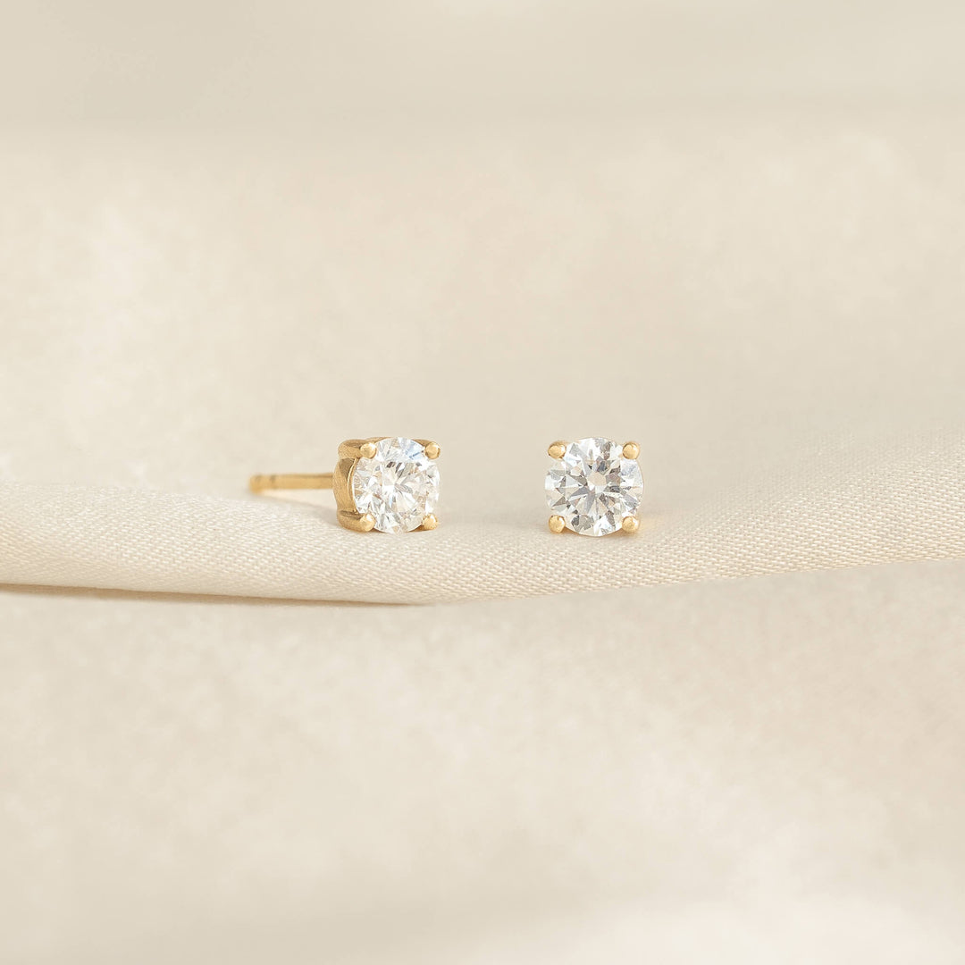 Two diamond stud earrings with gold settings are displayed on a soft, beige fabric. Their elegant sparkle conveys luxury and sophistication.