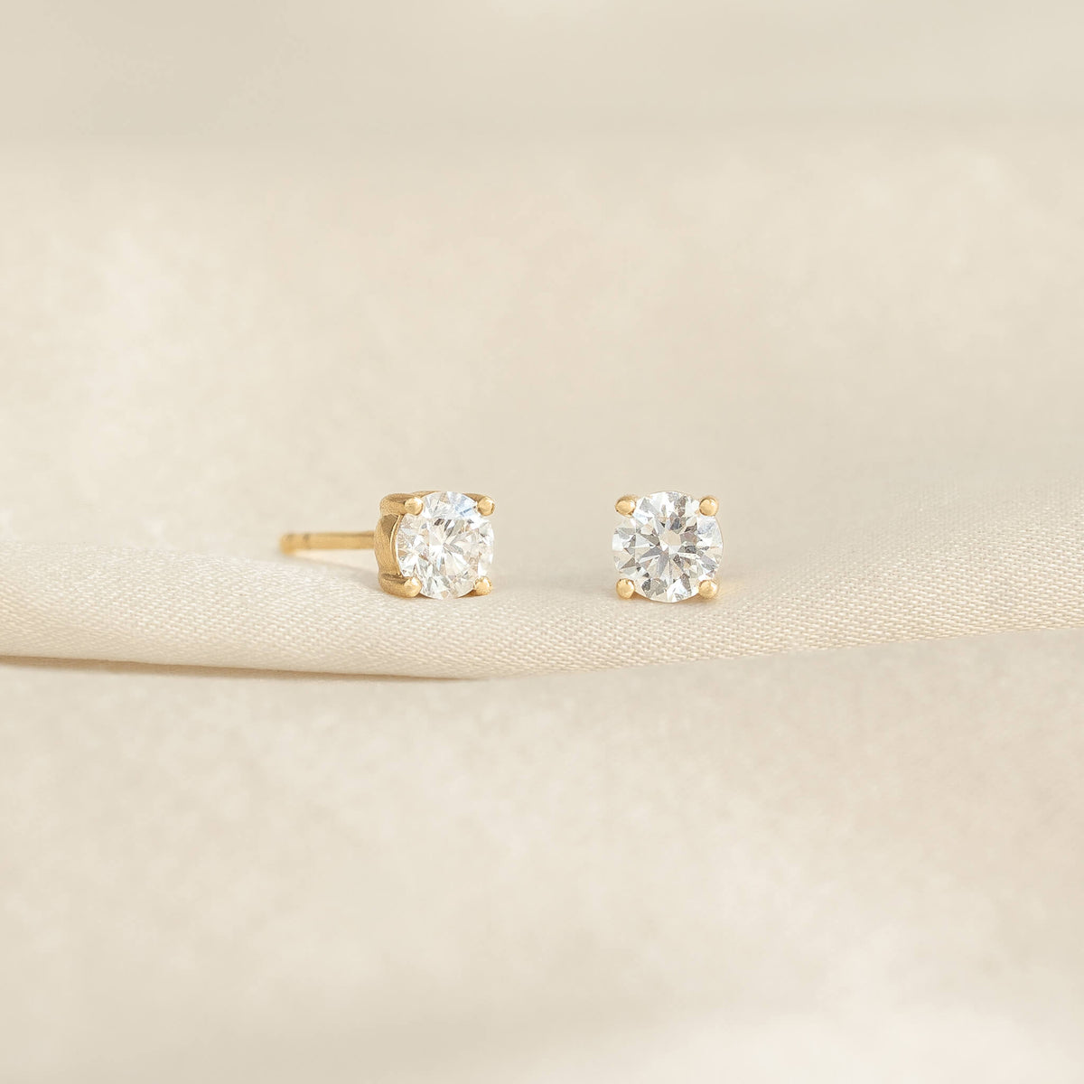 Two diamond stud earrings with gold settings are displayed on a soft, beige fabric. Their elegant sparkle conveys luxury and sophistication.