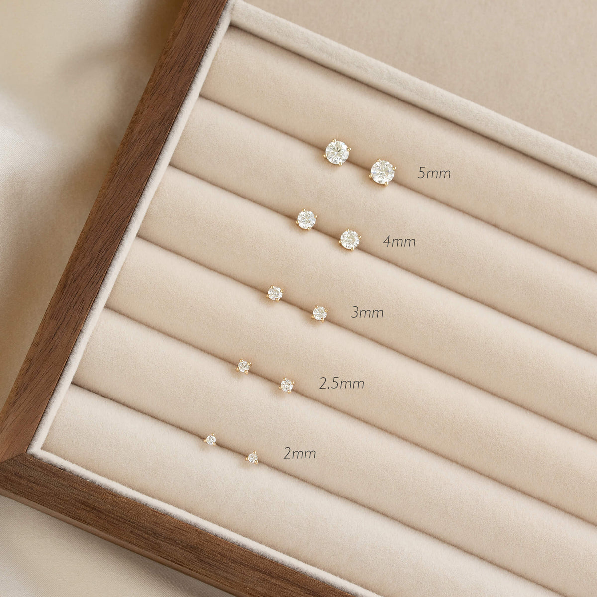 Elegant Gold jewelry display showing a set of diamond earrings, arranged by size from 2mm to 5mm, on a beige velvet holder with wooden frame.