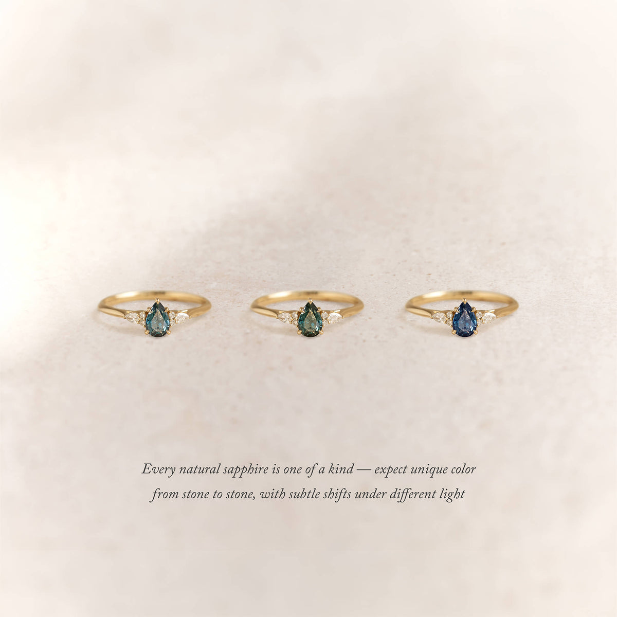 Three Gold rings with pear-shaped sapphires in varying hues are arranged side by side on a soft beige background.