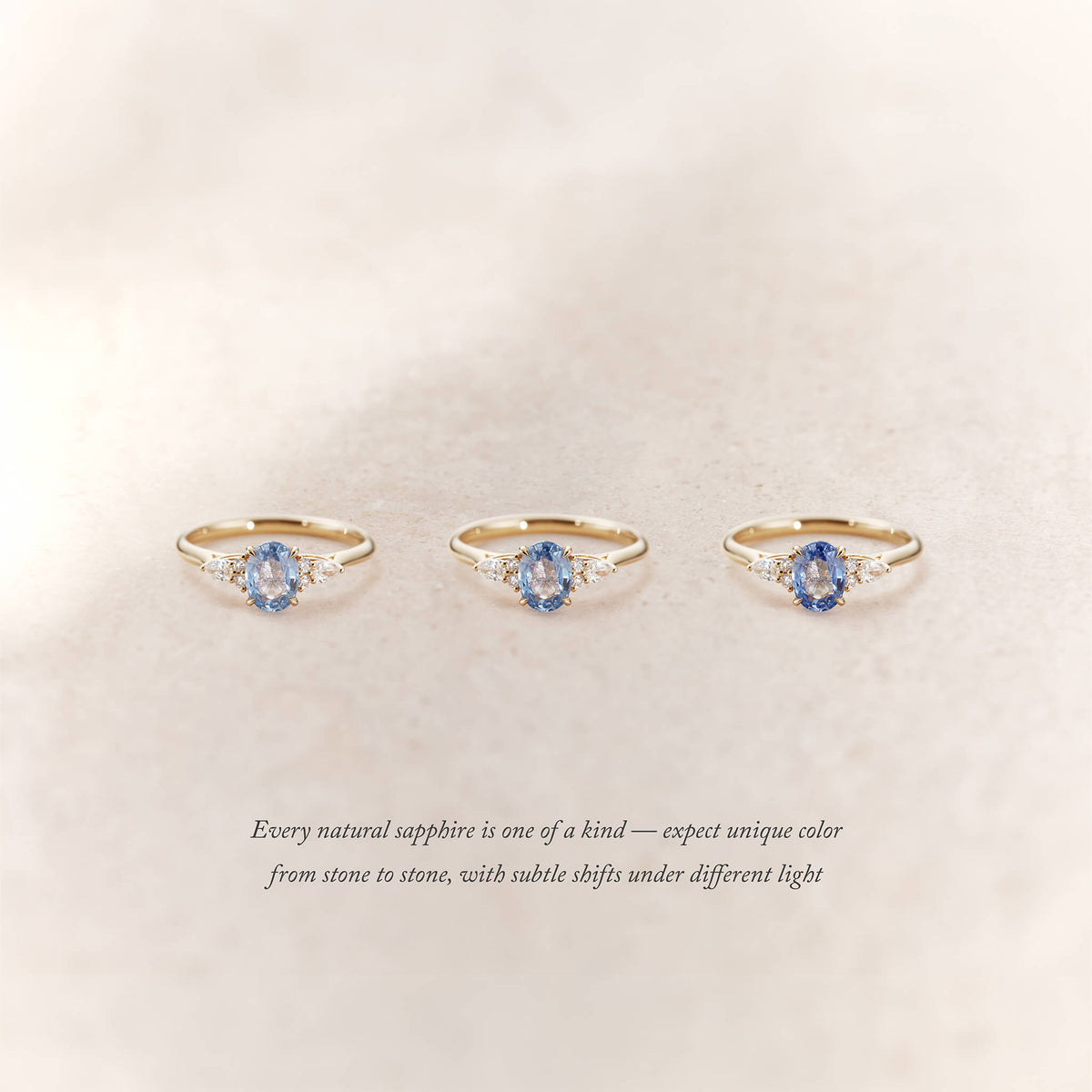 Three Yellow Gold rings with oval light blue sapphires and small diamond accents on each side are displayed side by side on a soft beige background.