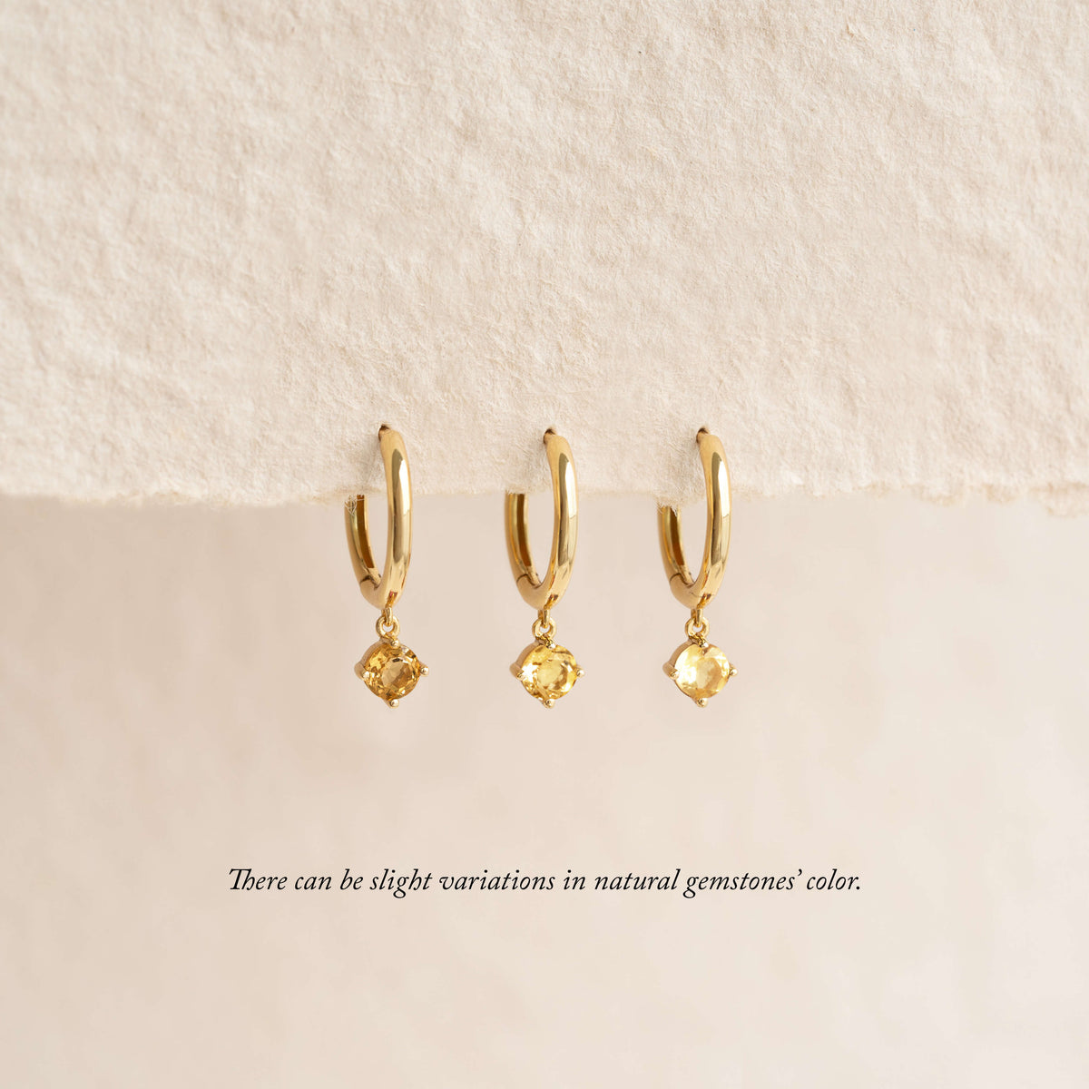Three gold hoop earrings with small dangling gemstones are displayed hanging from a soft, textured fabric background, with a note below indicating that natural gemstone colors may vary.