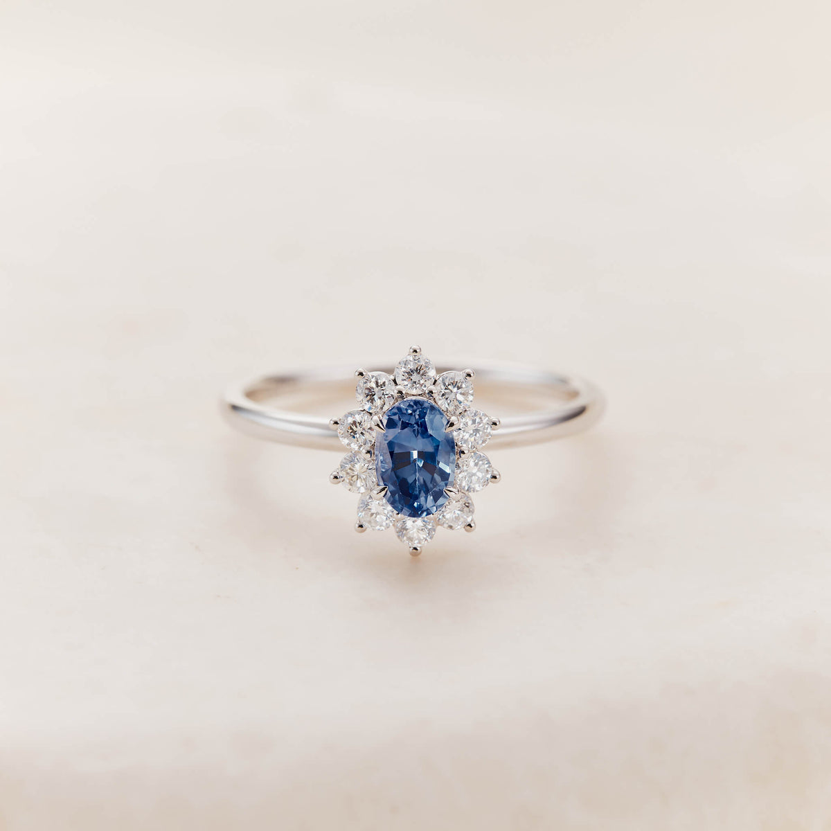 A White Gold halo ring featuring an oval-cut, deep blue sapphire center stone secured by four white gold prongs.