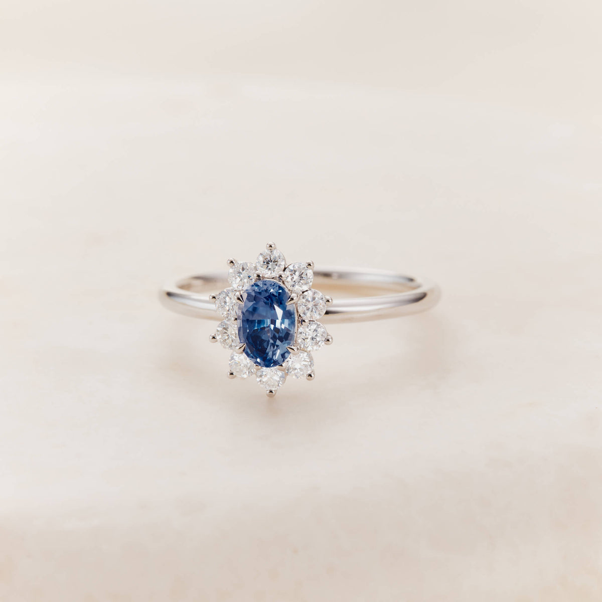 A White Gold halo ring featuring an oval-cut, deep blue sapphire center stone secured by four prongs.