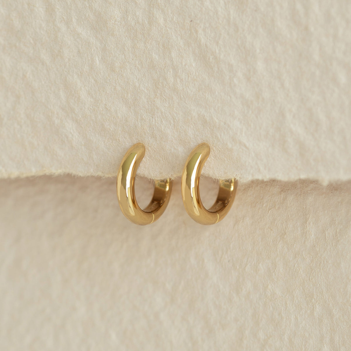 Chunky Huggie Hoop Earrings - Dione in gold vermeil displayed on soft beige textured background