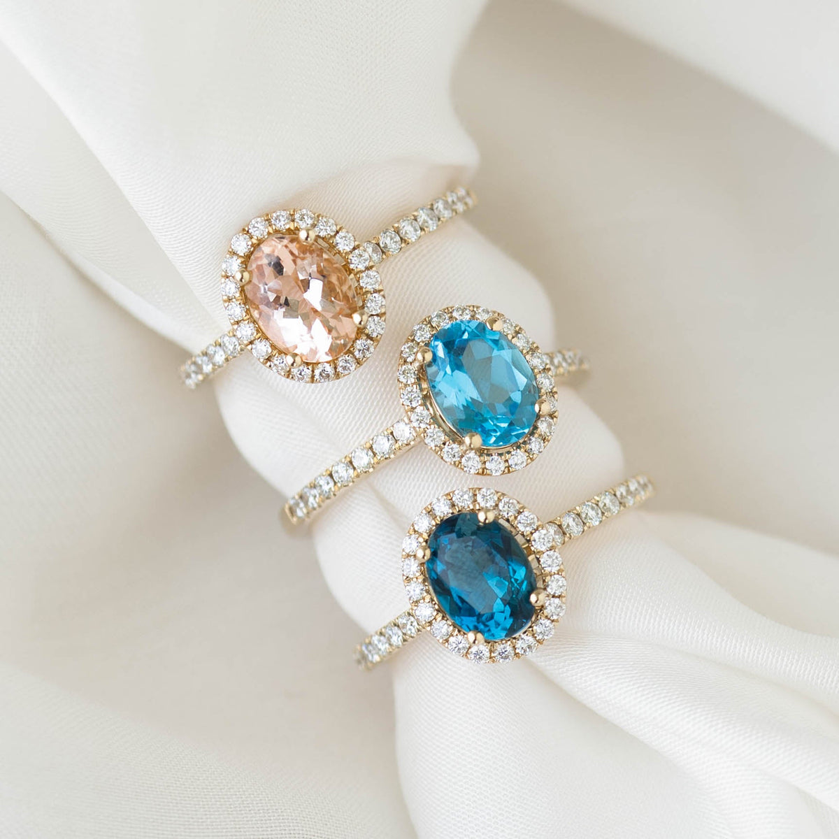 Oval Morganite Halo Ring Rose Gold - Suna displayed with two other oval halo rings on a soft white fabric background