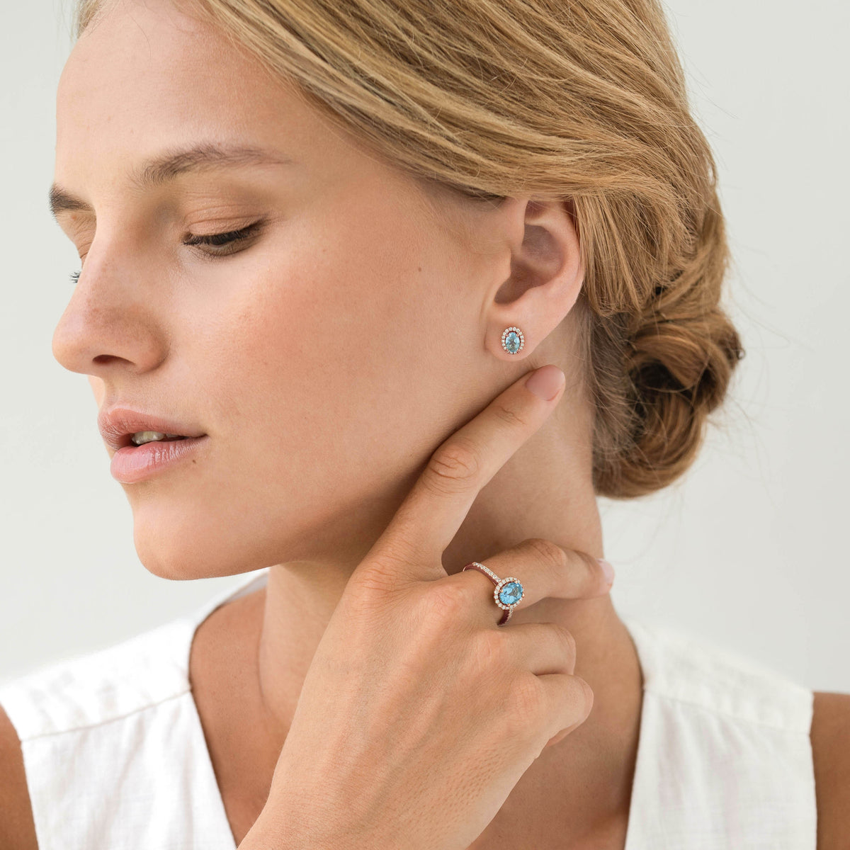 Close-up of woman wearing Oval Swiss Blue Topaz Halo Ring Rose Gold - Suna with matching earrings on a white top