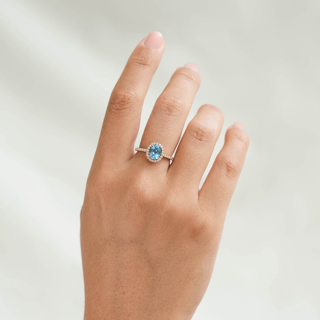 Close-up of a hand wearing an Oval Swiss Blue Topaz Halo Ring Rose Gold - Suna with diamond pavé band on a neutral background