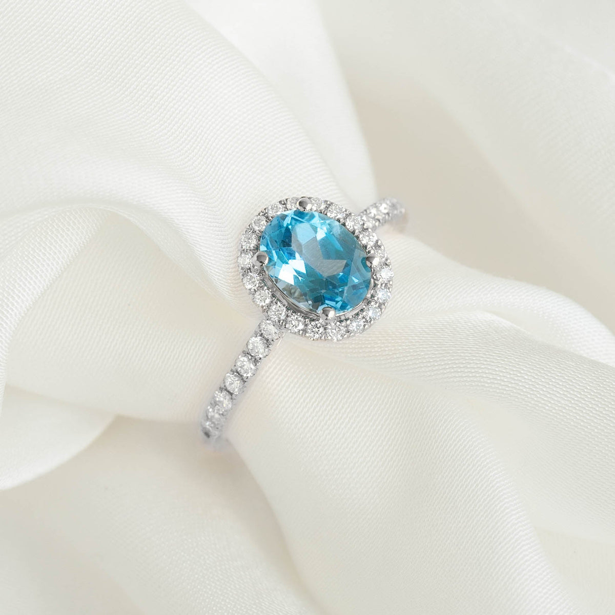 Oval Swiss Blue Topaz Halo Ring White Gold - Suna displayed on soft white fabric background with sparkling pavé diamonds