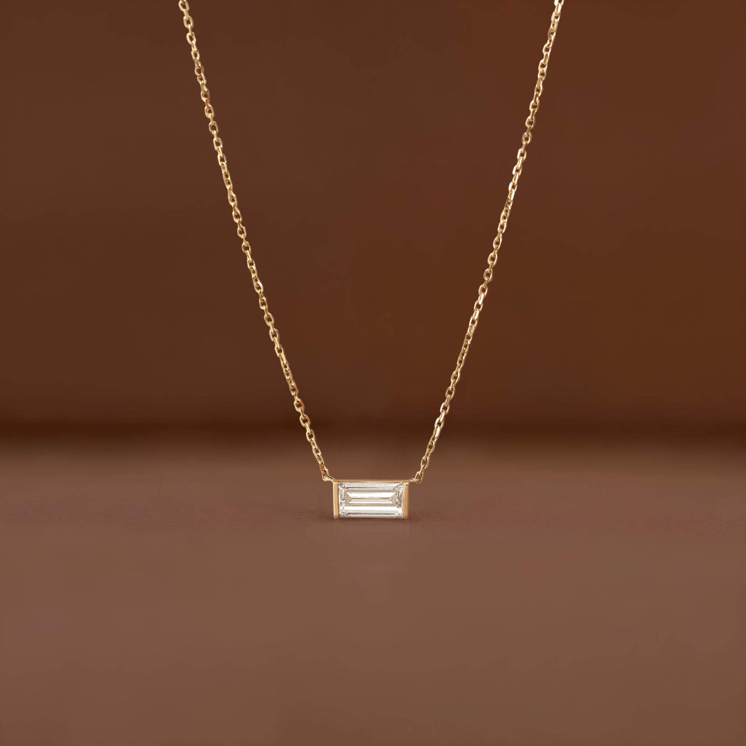 Delicate Gold chain necklace featuring a small rectangular baguette-cut clear gemstone pendant, displayed against a warm brown background.