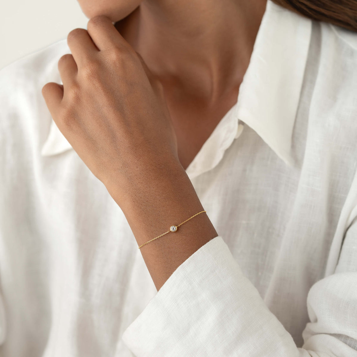 Close-up of a person wearing a white linen blouse, gently resting one hand on her chin while showcasing a delicate Gold Vermeil bracelet with a small White topaz gemstone centerpiece.