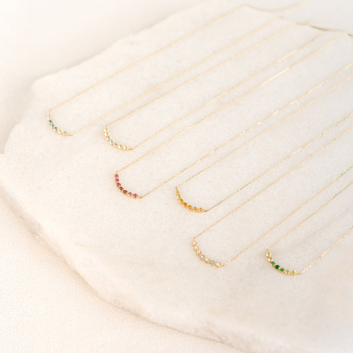 Close-up of six April Birthstone Necklace 14k Gold - Diamond pieces with colored stones on delicate gold chains on a beige surface
