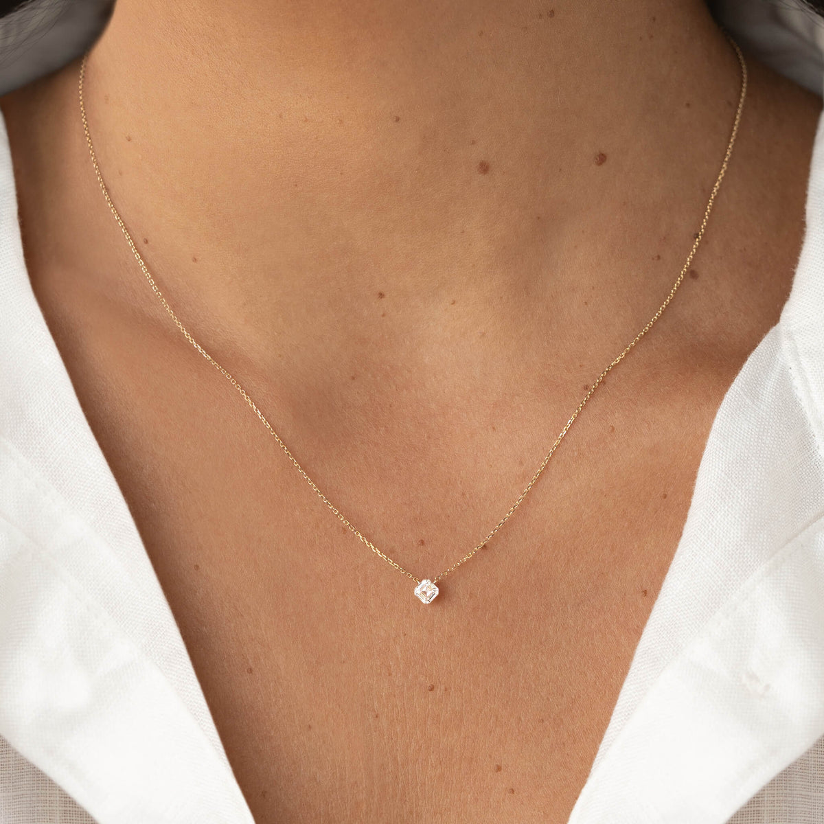 Close-up of a delicate Gold chain necklace with a small asscher-cut clear gemstone pendant, worn with a white V-neck blouse.