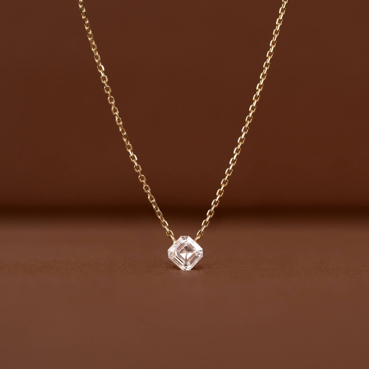 Close-up of a delicate Gold chain necklace featuring a clear asscher-cut gemstone pendant against a warm brown background.