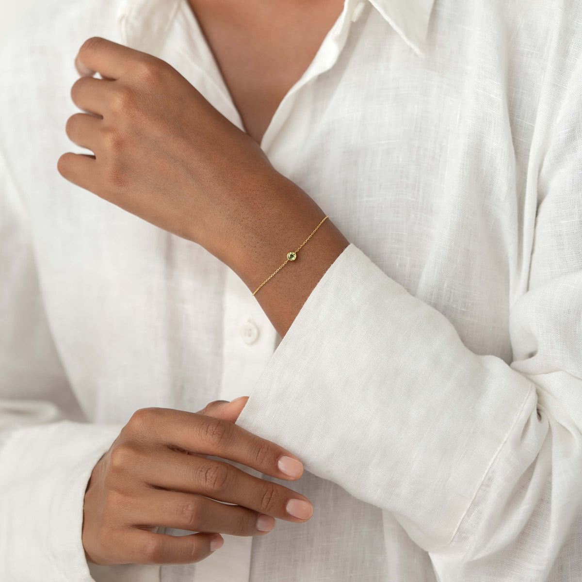 Close-up of a person wearing a white linen blouse, gently resting one hand while showcasing a delicate Gold Vermeil bracelet with a small Peridot gemstone centerpiece.