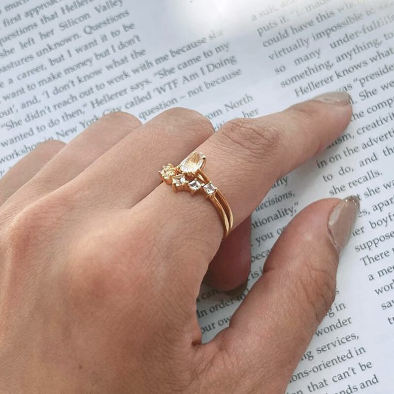 Close-up of a hand with neutral nail polish wearing two delicate gold rings with clear stones over an open book background