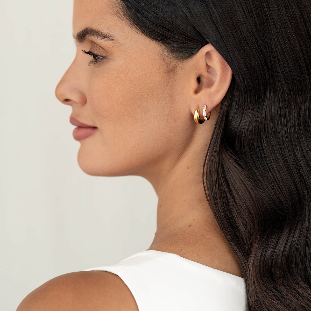 Side profile of a woman wearing two Gold Vermeil earrings—one plain and one set with baguette-cut gemstone—styled as an ear stack, shown against a soft neutral background with dark hair.