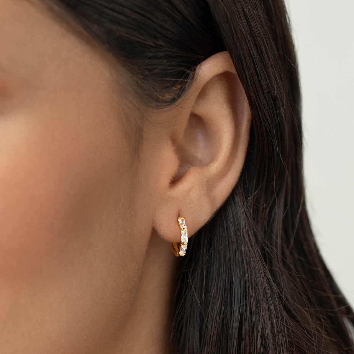 Close-up of a woman’s ear wearing a small Gold Vermeil earring set with baguette-cut gemstone, shown against a neutral background with dark hair tucked behind the ear.