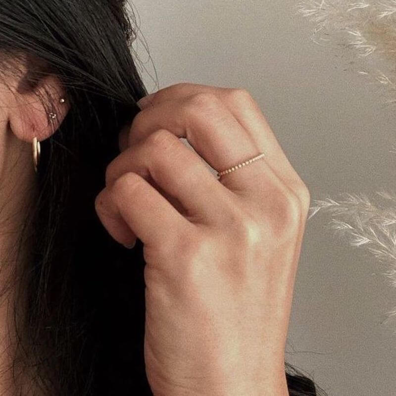 Close-up of a hand with a delicate beaded ring near the ear with small gold hoop and multiple ear piercings on a neutral background
