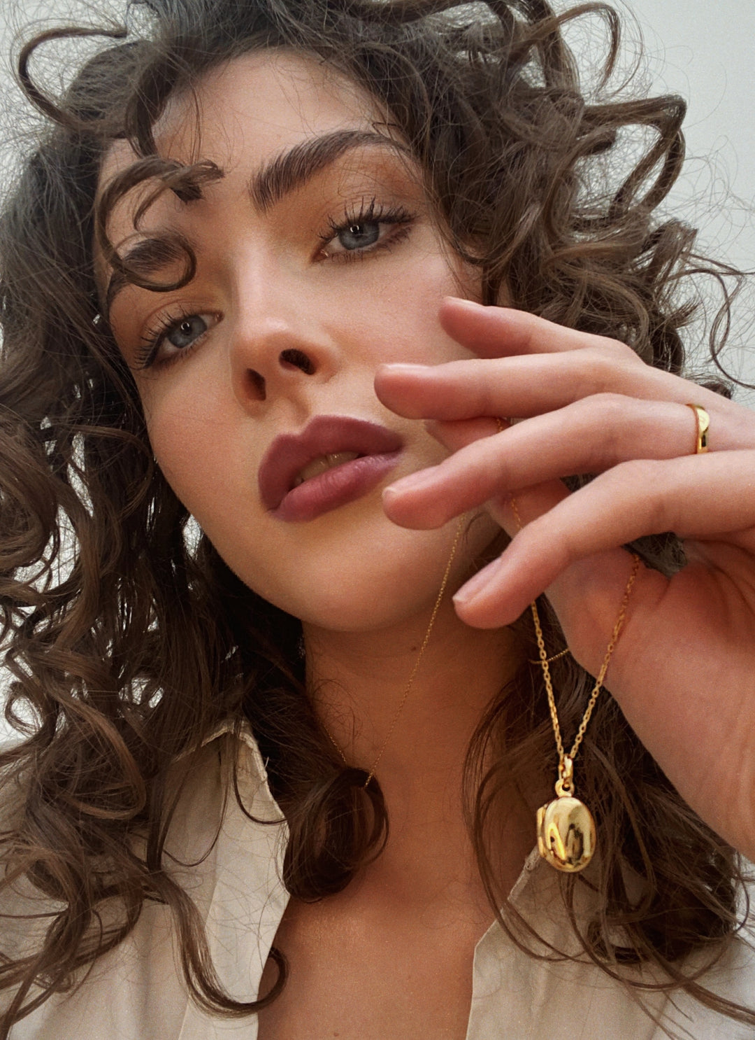 Close-up of a woman with curly hair holding a gold pendant necklace near her face against a white background