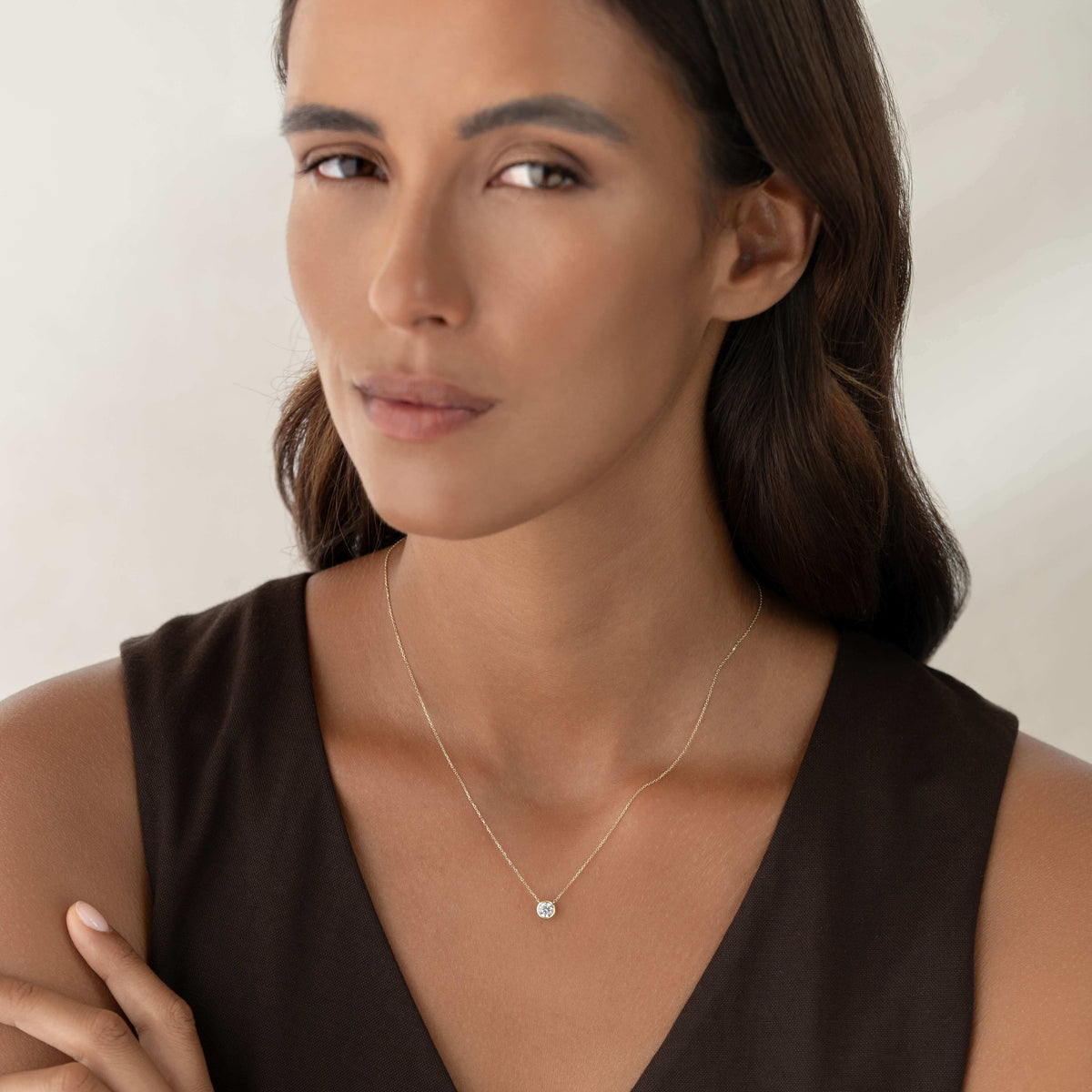 A portrait of a woman with dark hair wearing a delicate Gold chain necklace with a single round diamond solitaire pendant in a bezel setting, styled with a V-neck dark brown top.