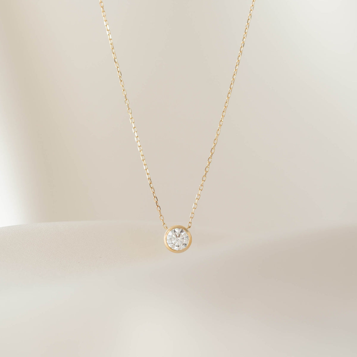 A close-up shot of a delicate Gold necklace featuring a round brilliant diamond solitaire pendant secured in a minimalist bezel setting, suspended elegantly against a soft, cream-colored fabric background.