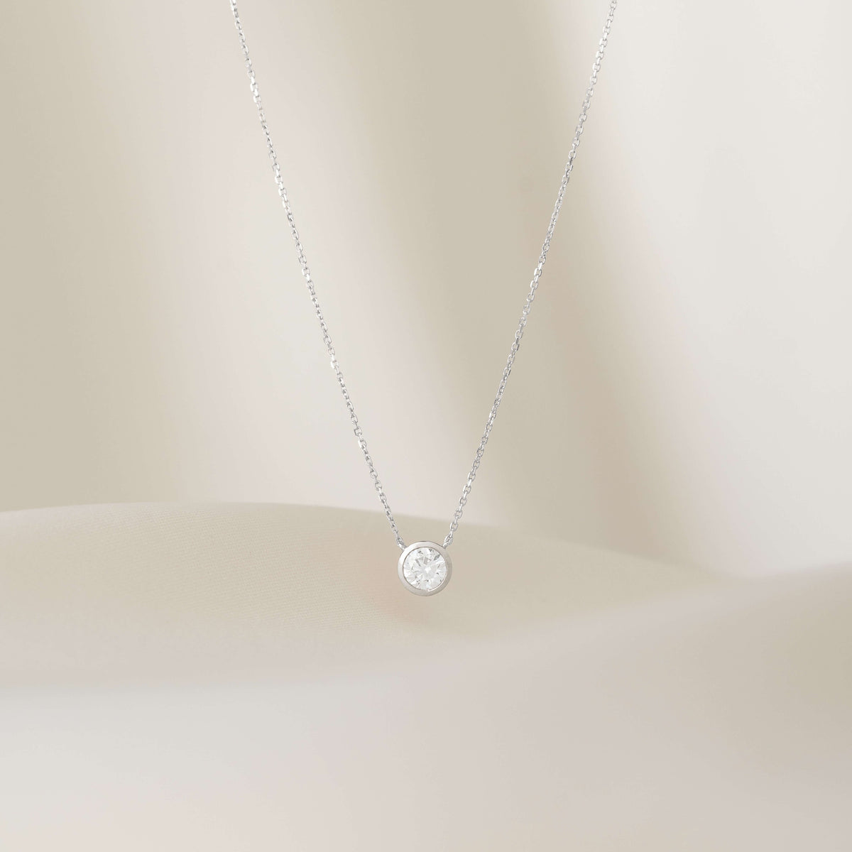A close-up shot of a delicate White Gold necklace featuring a round brilliant diamond solitaire pendant in a minimalist bezel setting, suspended against a soft, off-white background.