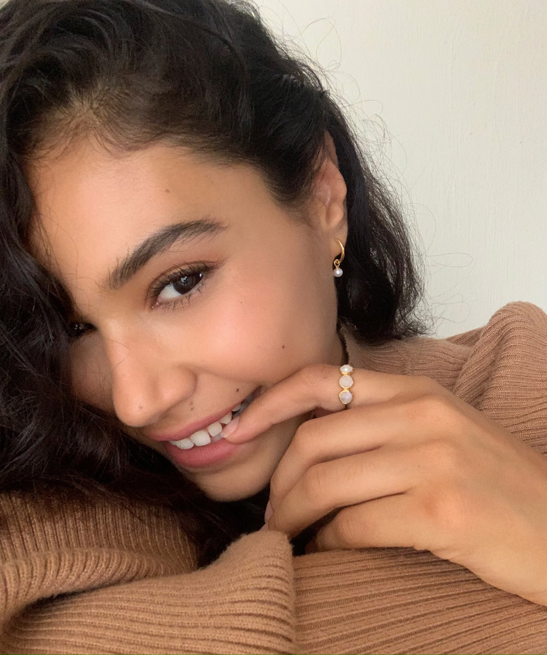 Close-up of a smiling woman with curly hair wearing a brown sweater, gold earrings, and a ring with white stones
