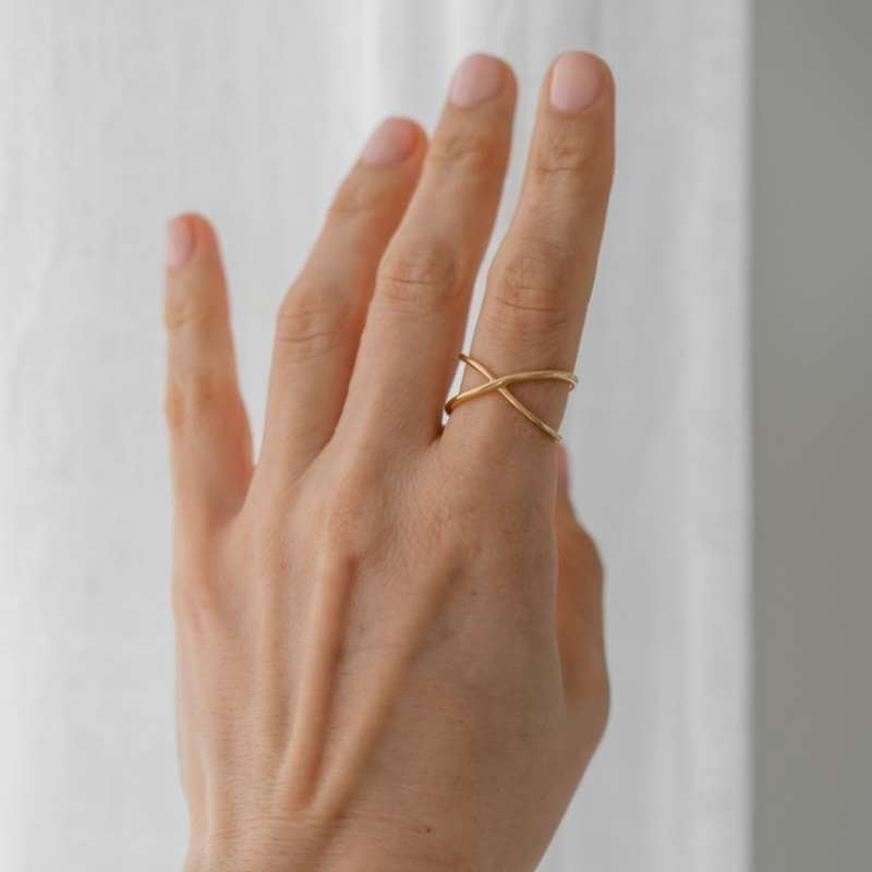Close-up of a hand wearing a delicate gold crisscross ring against a soft white background
