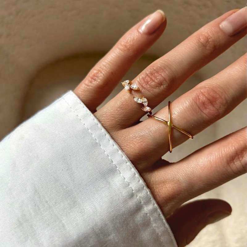 Close-up of a hand wearing two delicate rings, one with small clear stones and one with a gold X design against a neutral background