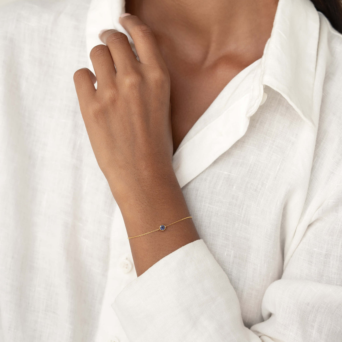 Close-up of a person wearing a white linen blouse, gently resting one hand while showcasing a delicate Gold Vermeil bracelet with a small Cordierite gemstone centerpiece.