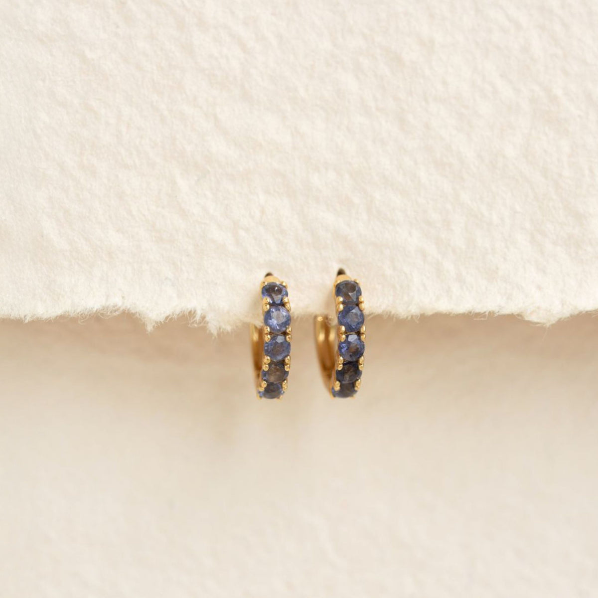 December Birthstone Huggie Earrings - Cordierite featuring gold vermeil and vibrant blue cordierite gemstones on a cream background