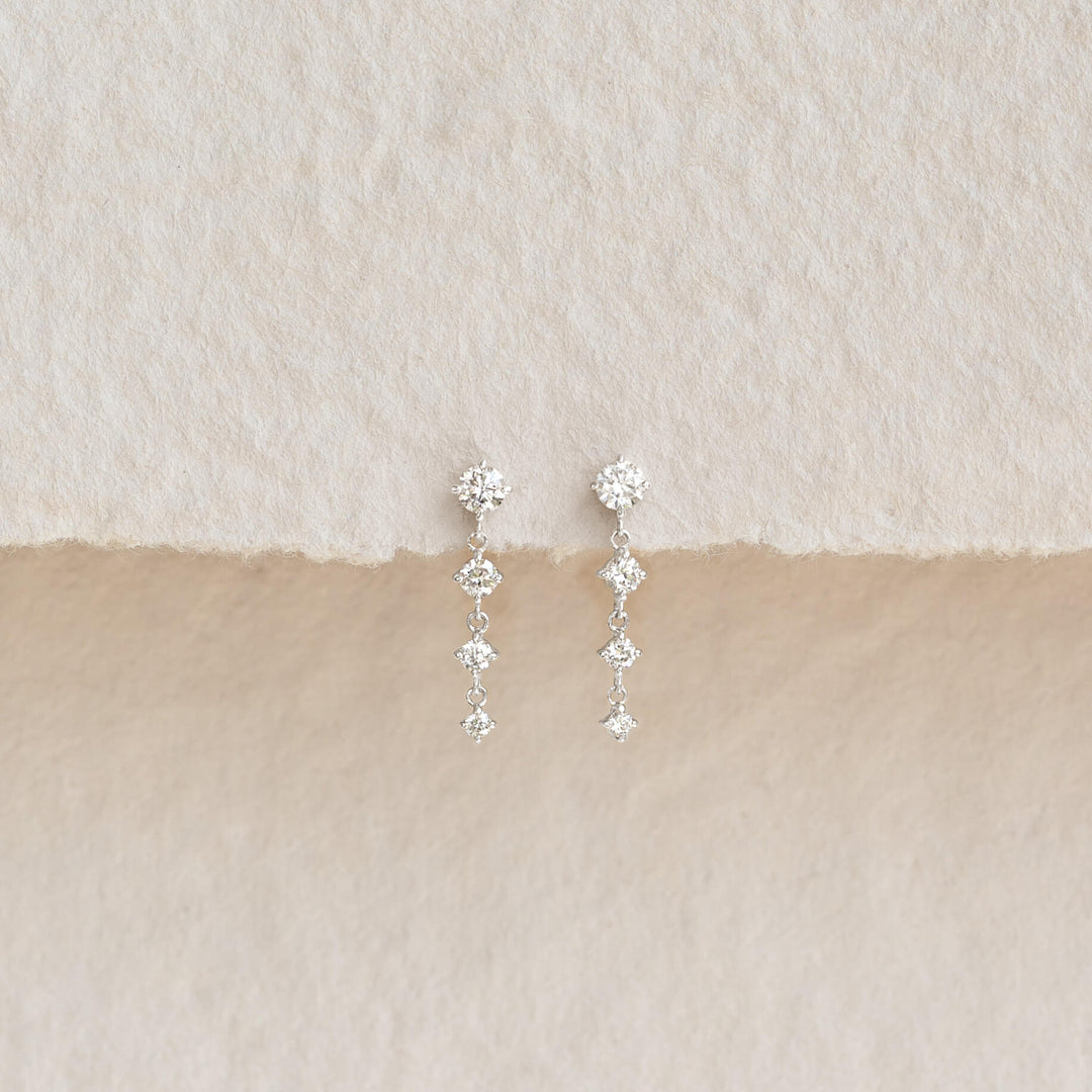 Pair of delicate drop earrings featuring sparkling diamond-shaped stones arranged in a vertical design, displayed on a textured beige and cream background.