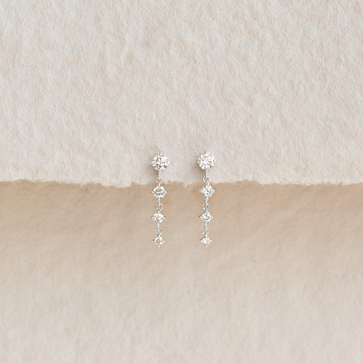 Pair of delicate drop earrings featuring sparkling diamond-shaped stones arranged in a vertical design, displayed on a textured beige and cream background.