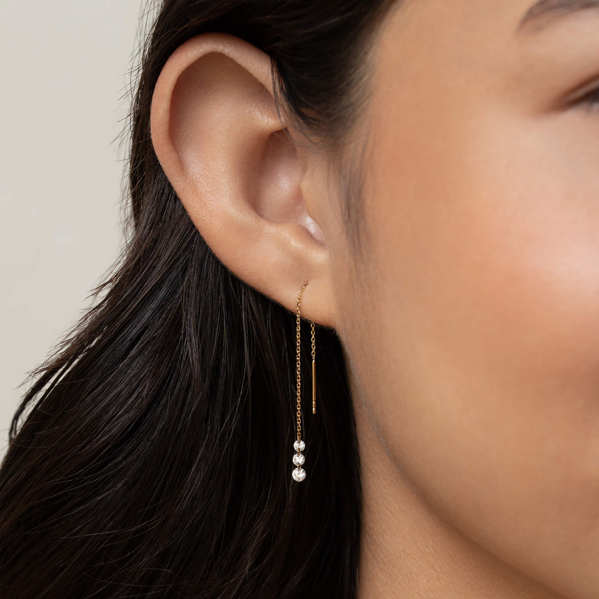 Close-up of a woman's ear wearing a delicate Gold chain earring, with three small sparkling gems hanging. Her dark hair and smooth skin add a soft elegance.