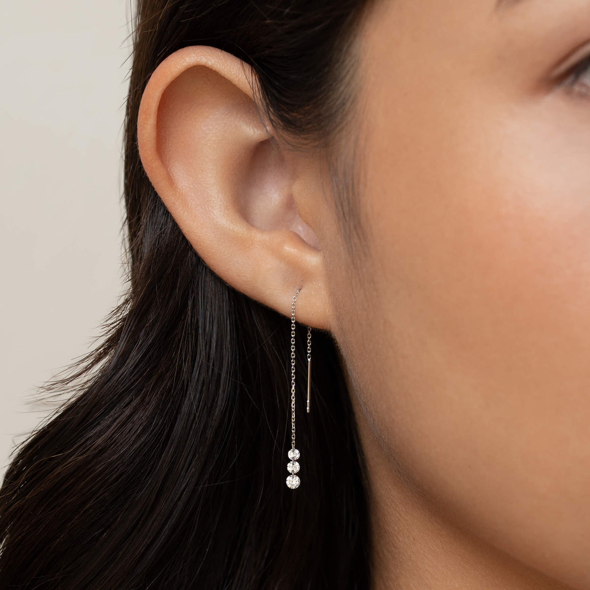 Close-up of a person’s ear wearing a delicate White Gold chain earring with three small, sparkling crystals. The tone is elegant and minimalistic.