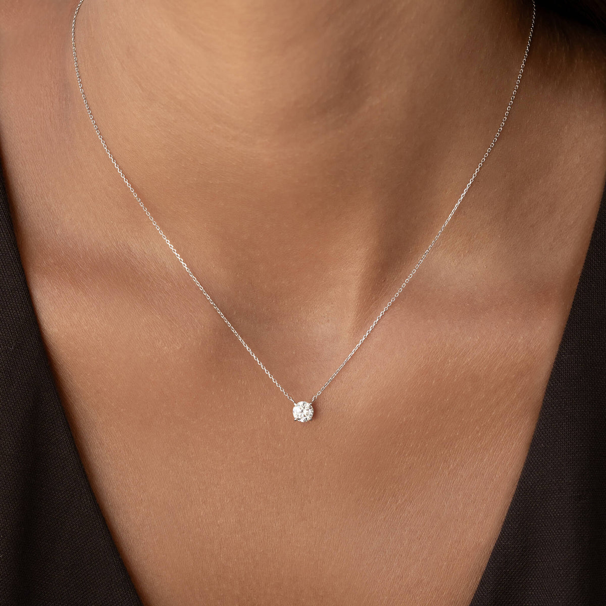 Close-up of a person’s neck wearing a delicate White Gold chain necklace with a single round diamond pendant centered at the collarbone, against a dark top background.