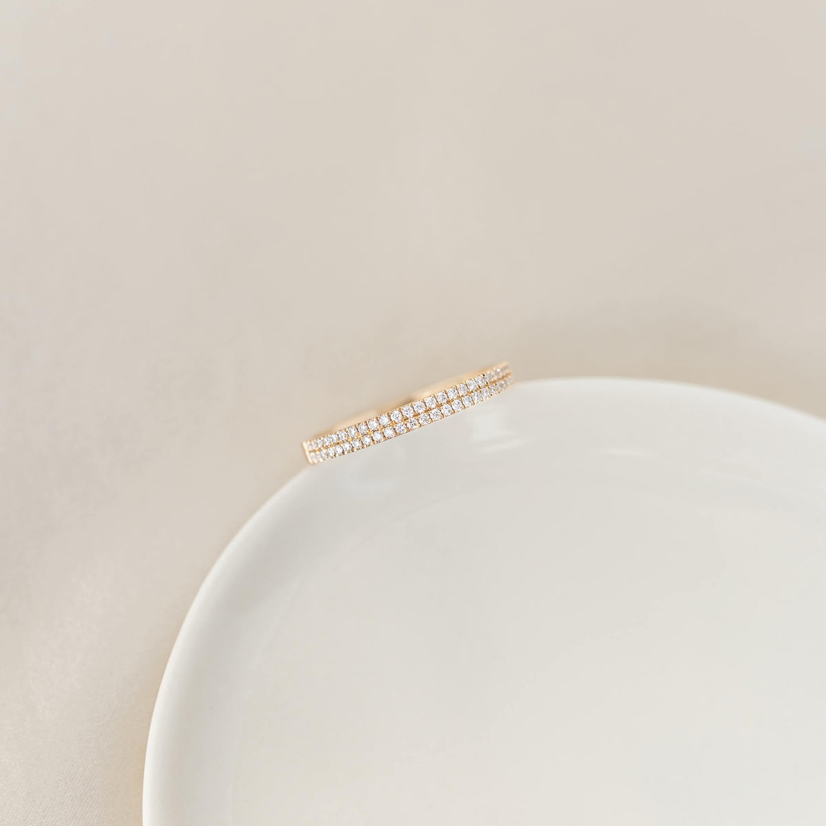 Double band half Gold ring with small sparkling diamonds, delicately balanced on the edge of a white ceramic plate against a soft warm neutral background.