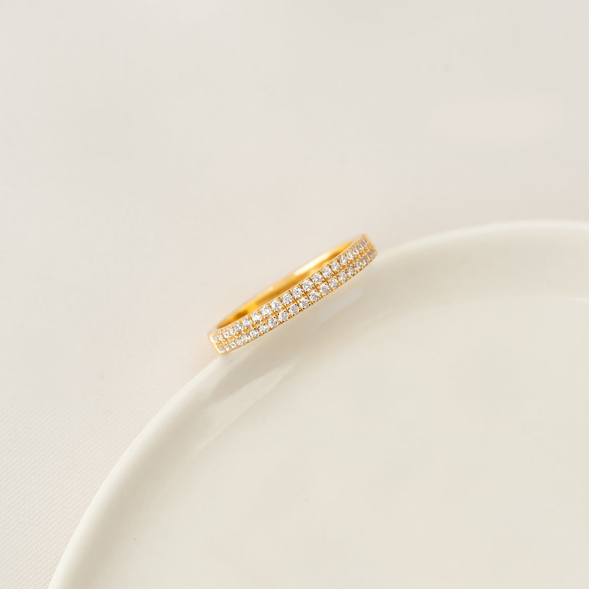 Gold band ring resting on the edge of a white ceramic dish against a soft, neutral background.