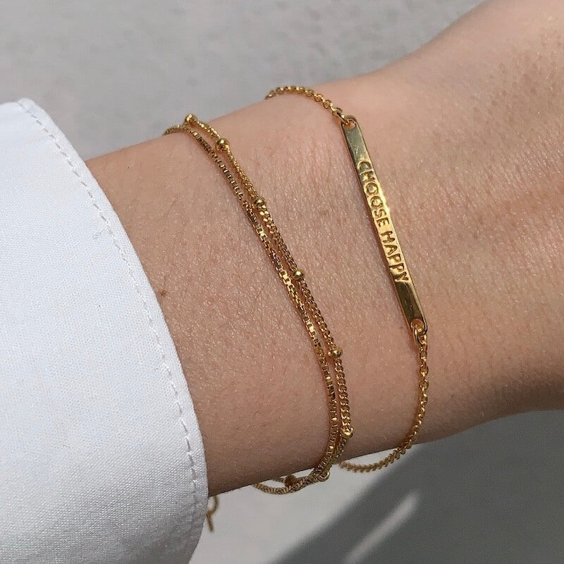 Close-up of a wrist with two delicate gold chain bracelets, one featuring a rectangular bar with the words Choose Happy