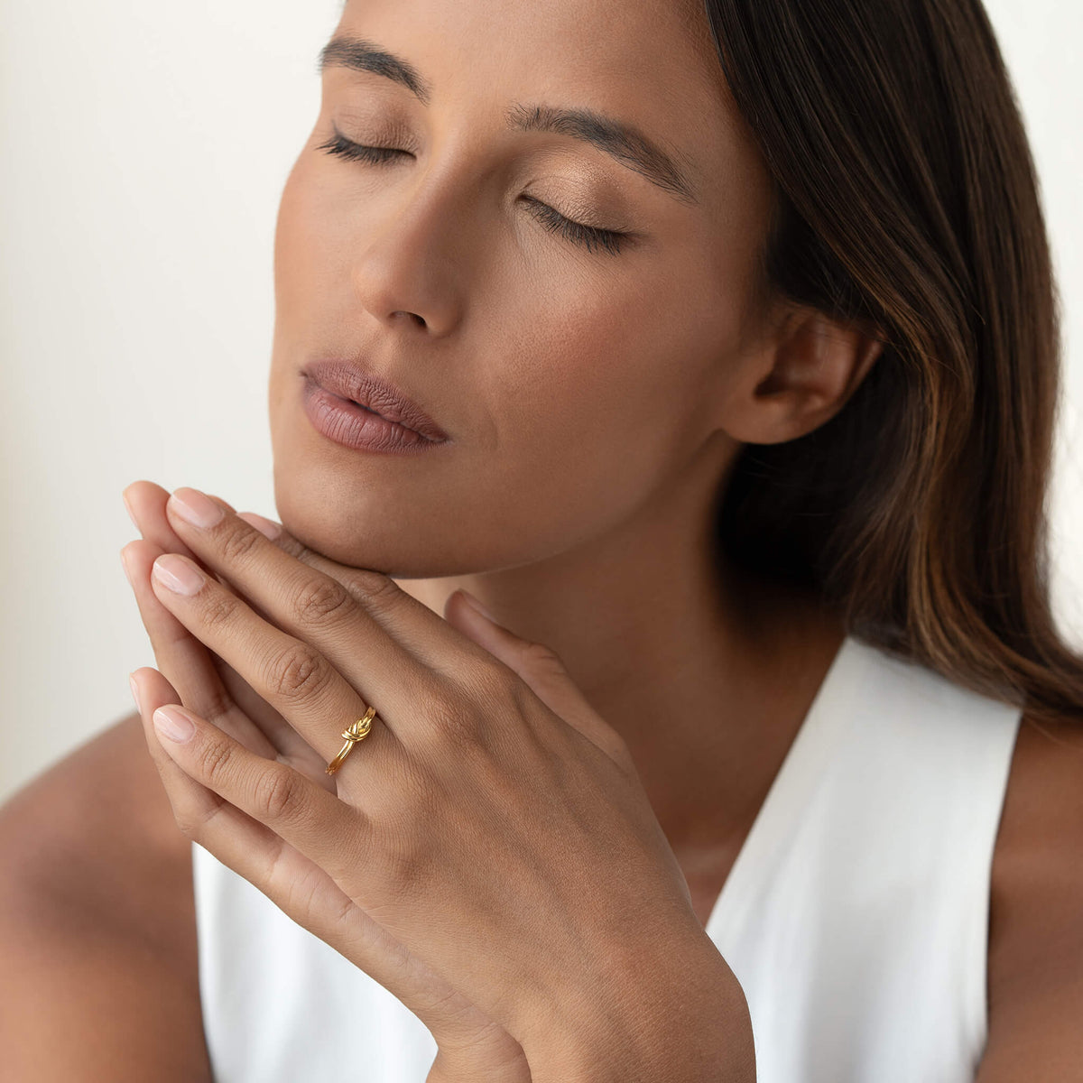 Portrait of a woman in a white sleeveless top gently resting her hands near her face while wearing a delicate Gold knot ring.