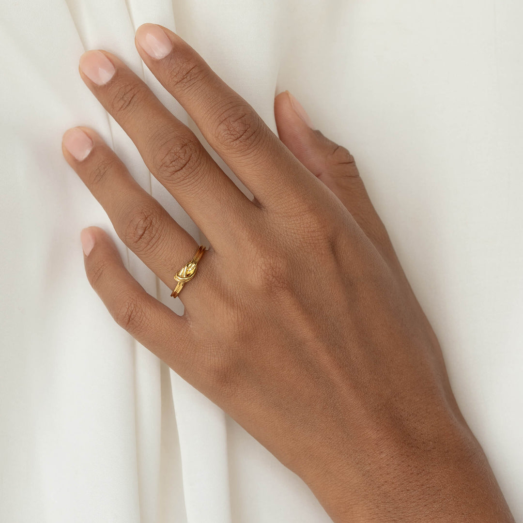 Close-up of a hand wearing a delicate Gold knot ring against softly draped white fabric.