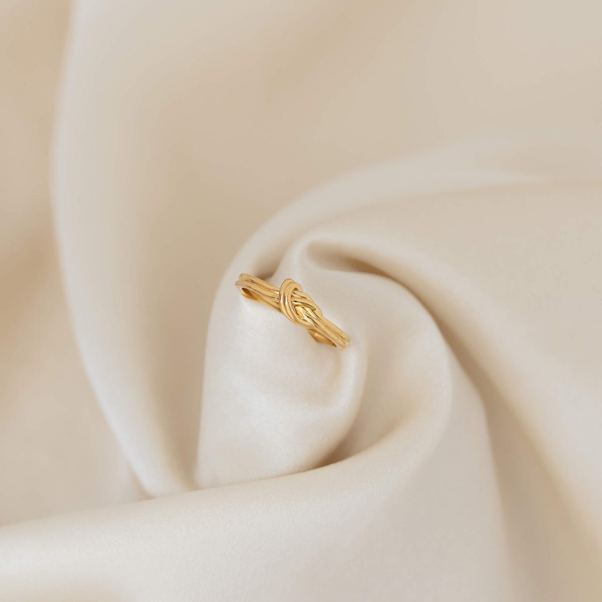 Delicate Gold knot ring displayed on softly folded cream satin fabric.