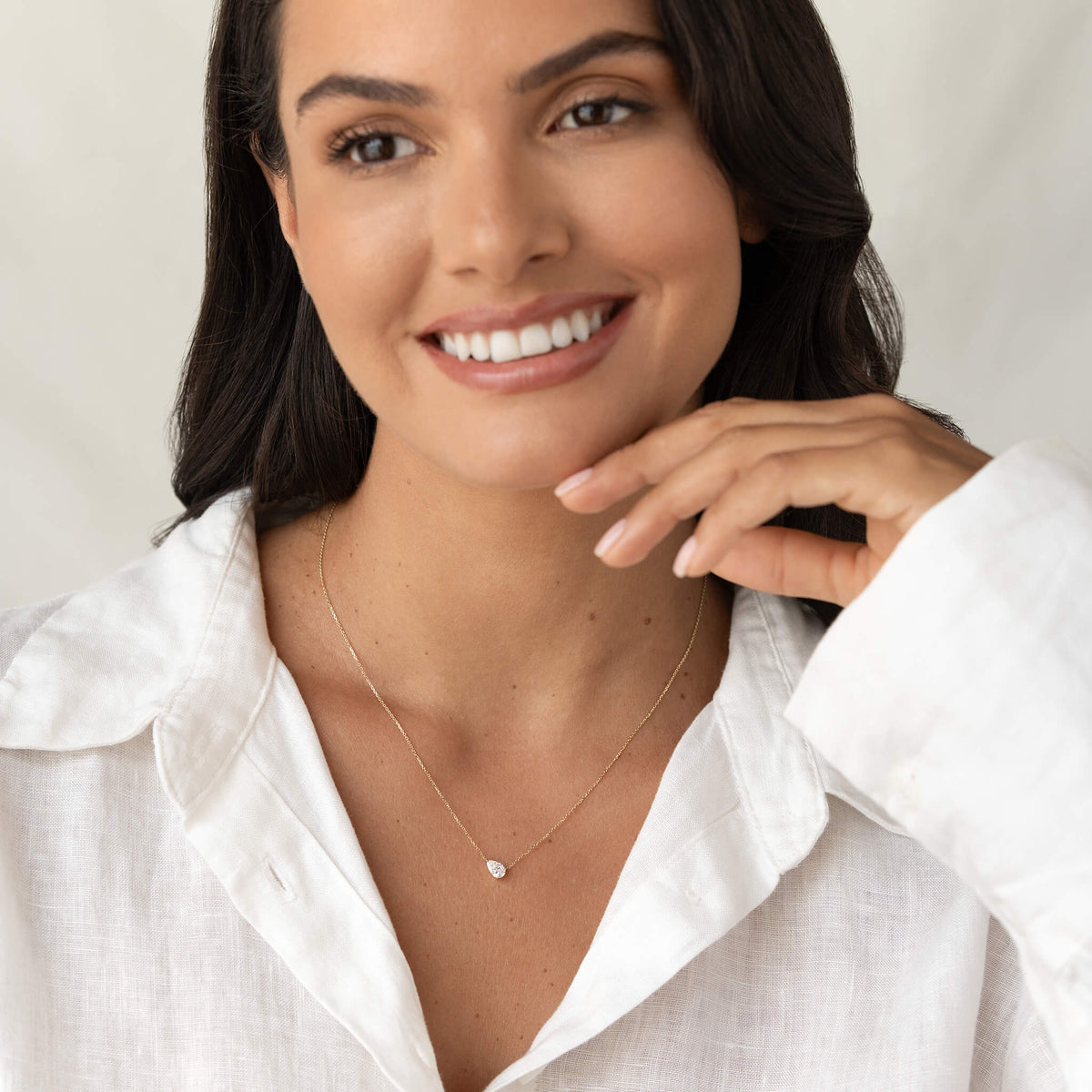 Portrait of a smiling woman in a white blouse showcasing a fine Gold necklace with a small pear-shaped gemstone pendant.