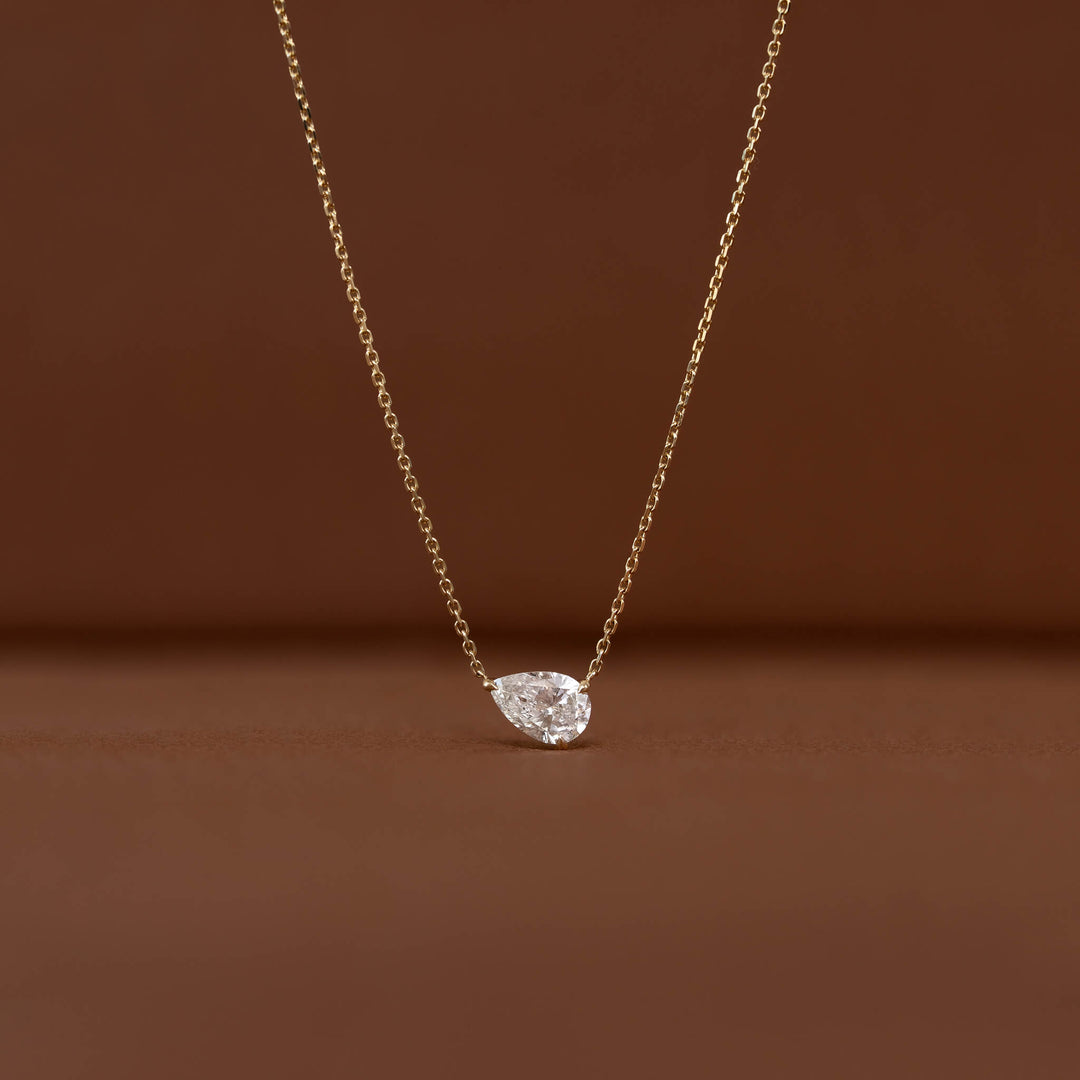 Close-up of a Gold chain necklace with a brilliant pear-shaped clear gemstone pendant displayed against a warm brown background.
