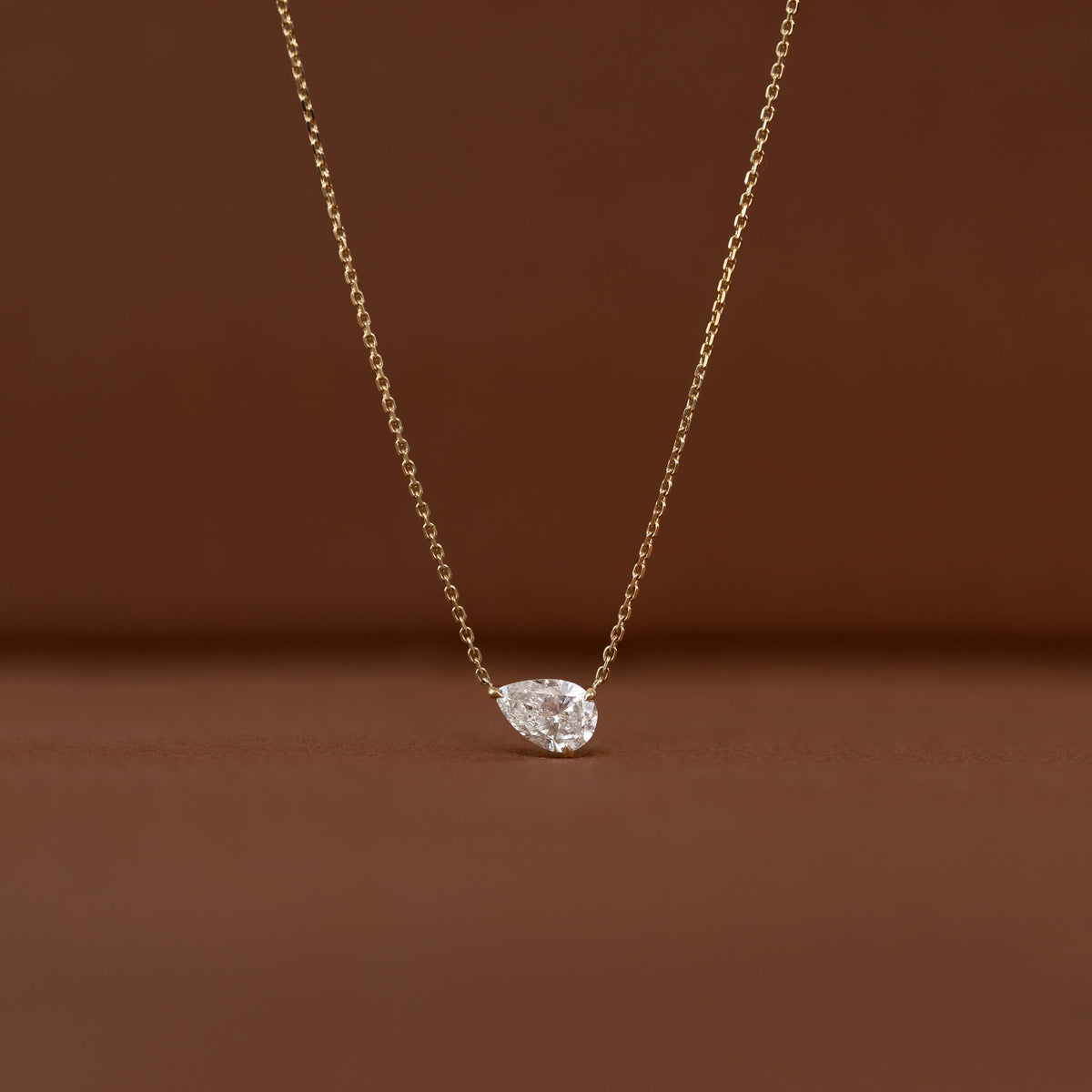 Close-up of a Gold chain necklace with a brilliant pear-shaped clear gemstone pendant displayed against a warm brown background.