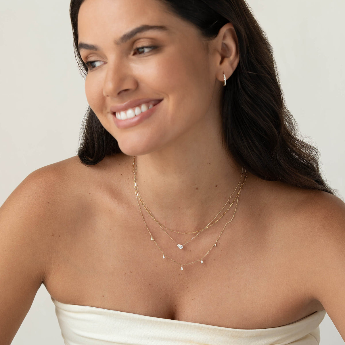 Smiling woman in a strapless white top wearing layered Gold necklaces with delicate gemstone accents and matching hoop earrings.