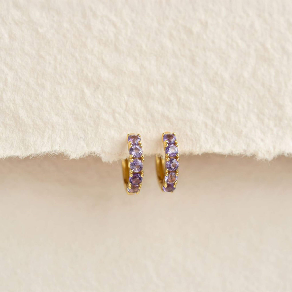 February Birthstone Huggie Earrings - Amethyst with gold vermeil setting on a soft beige textured background